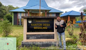 Enumerator survei publik berfoto di depan plang desa lokasi pengumpulan data lapangan